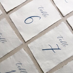 以下が含まれることがあります： 白い紙に青いインクで手書きの書体で書かれたテーブル番号カードのセット。番号は5、6、7、11、1です。各カードには、番号の上に「テーブル」という言葉が書かれています。