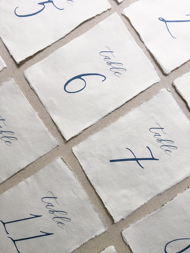以下が含まれることがあります： 白い紙に青いインクで手書きの書体で書かれたテーブル番号カードのセット。番号は5、6、7、11、1です。各カードには、番号の上に「テーブル」という言葉が書かれています。