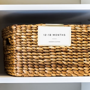 May include: A woven wicker basket with a label that reads "12-18 Months Hudson's Clothes".