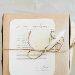 May include: A brown box with a white card attached to it. The card has the text "Welcome to Joyce's Bridal Shower" and a schedule of events. The box is tied with twine and decorated with dried flowers.