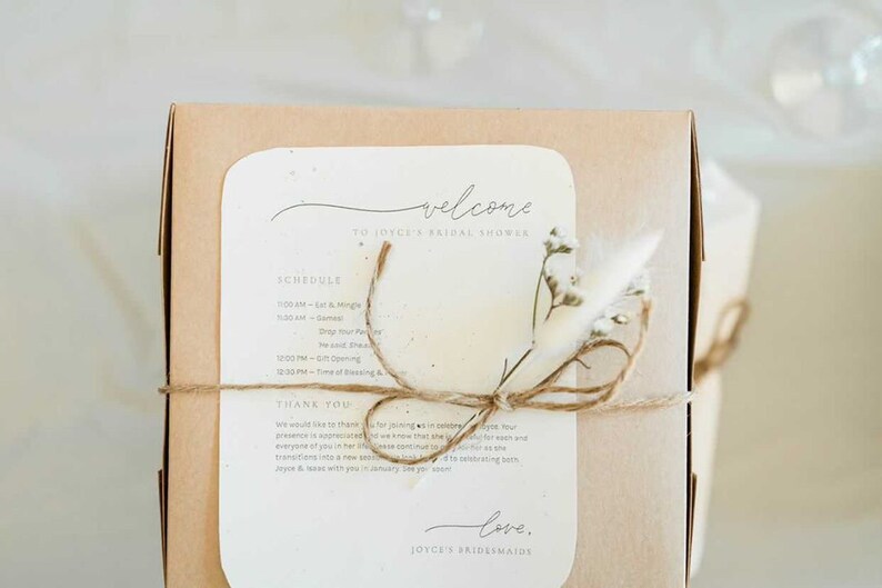May include: A brown box with a white card attached to it. The card has the text "Welcome to Joyce's Bridal Shower" and a schedule of events. The box is tied with twine and decorated with dried flowers.