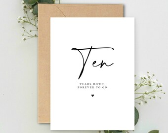 May include: A white card with the word "Ten" in elegant script, accompanied by the text "Years down, forever to go" and a small heart. The card is placed in a brown envelope and decorated with sprigs of white flowers and greenery.