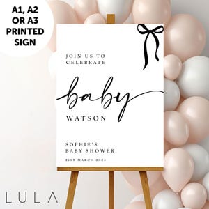 May include: A white printed sign on a wooden easel, with the text "baby Watson" in elegant script, and "Sophie's Baby Shower 21st March 2026". The sign is decorated with a black bow and surrounded by pink and white balloons.