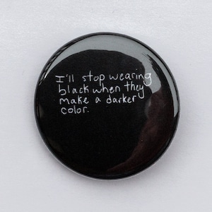 Puede incluir: Un botón negro brillante con el texto escrito a mano: "Dejaré de usar negro cuando hagan un color más oscuro". El botón tiene forma ovalada y está sobre una superficie blanca. Un accesorio humorístico.