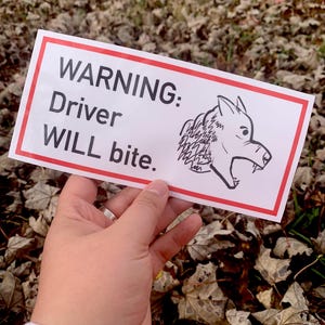 May include: A rectangular white sign with a red border, held by a hand. The sign reads "WARNING: Driver WILL bite." A black and white illustration of a wolf's head is on the right side of the sign.