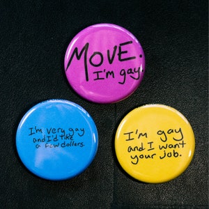May include: Three colorful round buttons with text. A purple button reads "MOVE. I'm gay." A blue button says "I'm very gay and I'd like a few dollars." A yellow button reads "I'm gay and I want your job."