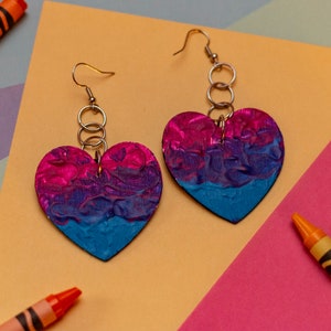 May include: A pair of heart-shaped earrings in pink, purple, and teal. The earrings hang from silver-coloured hooks and rings. The background includes crayons and colourful geometric shapes.
