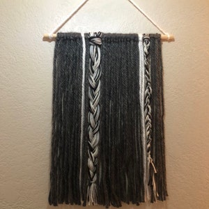 May include: A gray and white yarn wall hanging with a braided design. The hanging is attached to a wooden dowel rod and has a white string for hanging.