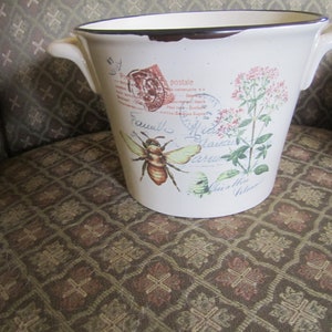 May include: White ceramic planter with brown rim and handles. The planter has a floral design with a bee and the text "Famille Fleurs" in cursive.