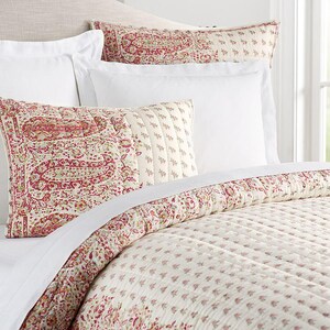 May include: Bedding set featuring a paisley patterned pillow and a floral patterned pillow. The quilt and pillows have a cream background with red and green accents. White sheets and a headboard complete the bed.