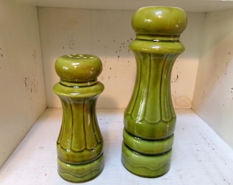 May include: Two green ceramic decorative items. One is a salt shaker with a rounded top and a series of holes. The other is a pepper grinder with a similar design, but taller. Both have a glossy finish and a ribbed texture.