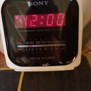 May include: A white Sony Dream Machine digital clock radio. The display shows 12:00 in red digits. It has FM/AM radio frequency options and the text "DREAM MACHINE" at the bottom.