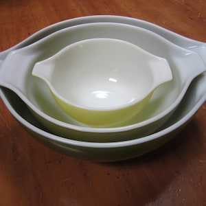 May include: A set of four nesting Pyrex bowls in shades of green and white. The bowls have a classic design with handles and a smooth, glossy finish. The largest bowl is approximately 10 inches in diameter.