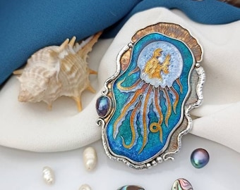 Pode incluir: Um broche de prata e cobre com um design de água-viva de esmalte azul e dourado. O broche é rodeado por pérolas brancas e conchas de abalone.