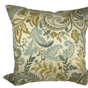 Könnte beinhalten: Quadratisches Zierkissen mit cremefarbenem Hintergrund und großem Blumenmuster in Blau-, Grün- und Goldtönen. Das Muster zeigt wirbelnde Blätter und florale Elemente. Das Kissen hat eine quadratische Form, ideal für Sofas.