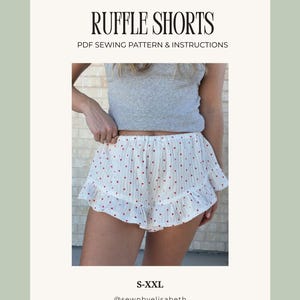 Op de afbeelding: Witte ruffle shorts met een rood hartjespatroon. De shorts hebben een gefranjerde zoom en maken deel uit van een PDF naaipatroon. De tekst "RUFFLE SHORTS" en "PDF SEWING PATTERN & INSTRUCTIONS" wordt weergegeven.