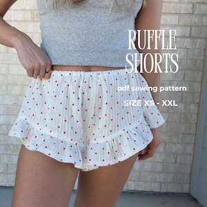 May include: White ruffle shorts with a red heart pattern. The shorts have a ruffled hem and are shown with a grey top. Text on the image reads "RUFFLE SHORTS" and "pdf sewing pattern" with size options.