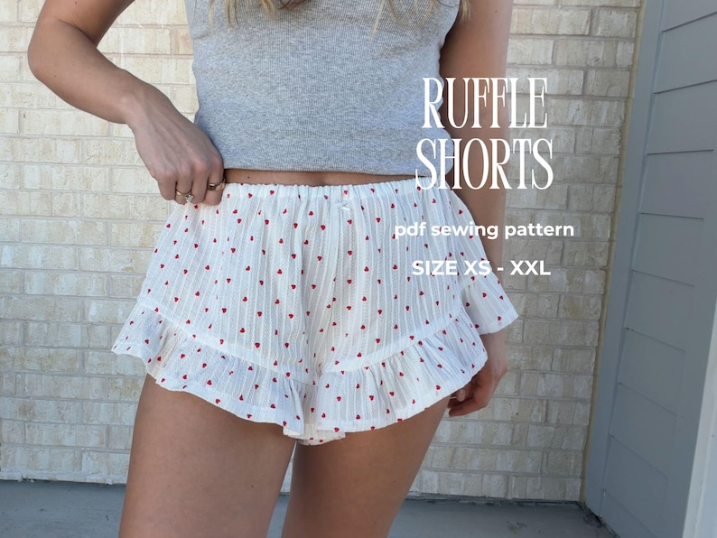 Op de afbeelding: Witte ruffle shorts met een rood hartjespatroon. De shorts hebben een gefranjerde zoom en worden getoond met een grijze top. Tekst op de afbeelding luidt "RUFFLE SHORTS" en "pdf sewing pattern" met maatopties.