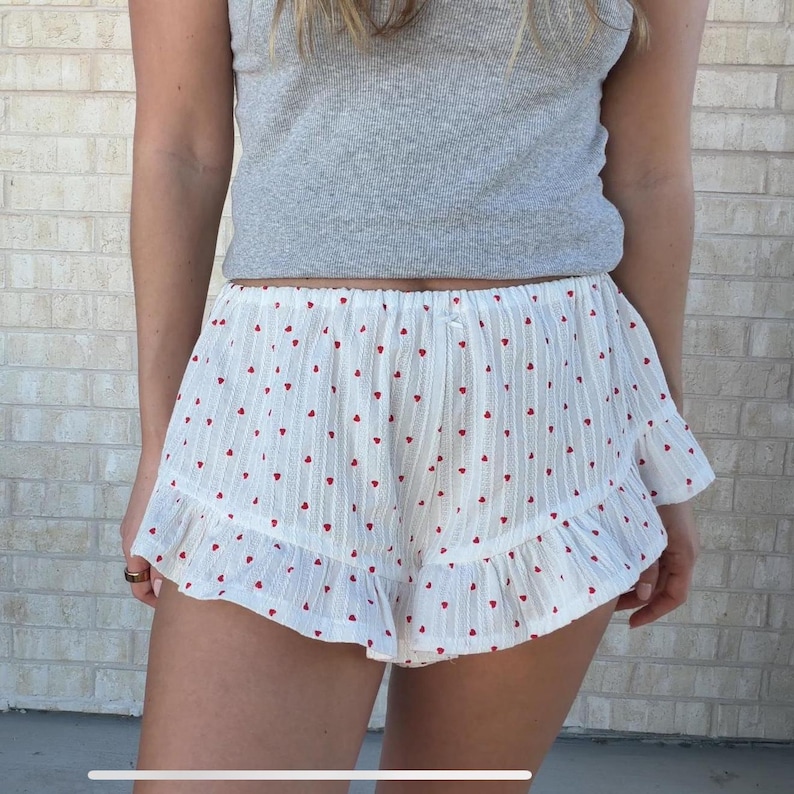 Op de afbeelding: Witte shorts met een patroon van kleine rode hartjes en een gefranjerde zoom. De shorts worden gecombineerd met een grijze tanktop. De shorts hebben een verticaal gestreepte textuur.