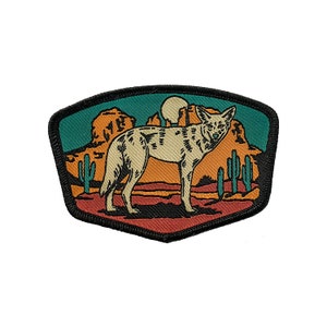May include: A coyote stands in a desert landscape with a sunset background. The coyote is beige with black markings. The desert landscape is a mix of orange, red, and green colors. There are cacti in the background.