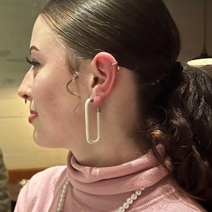 May include: A woman wearing a pink turtleneck sweater and a pearl necklace. She is also wearing white rectangular hoop earrings and has multiple ear piercings. Her hair is pulled back in a ponytail.