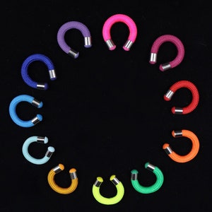 May include: A collection of colorful, horseshoe-shaped earrings arranged in a circle against a black background. The earrings are made of a thick, braided cord in various colors, including purple, pink, blue, red, orange, yellow, and green. Each earring has silver-colored metal ends.