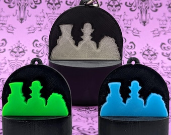 May include: Three black, rounded keychains featuring a raised silhouette of three figures in top hats. One keychain has a silver-coloured inlay, one has a bright green inlay, and one has a bright blue inlay. Each includes a metal ring for attachment.