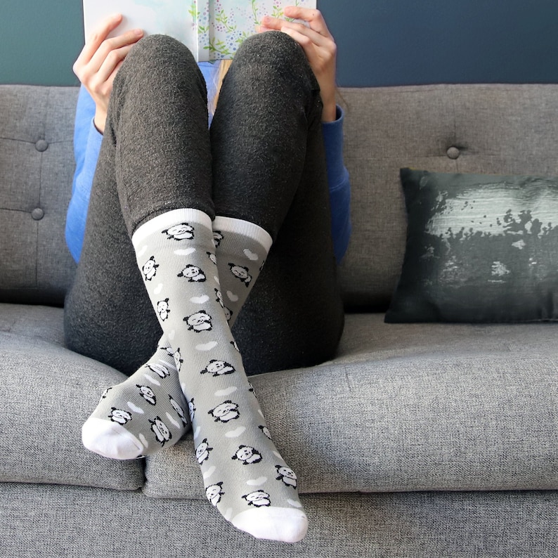 K&ouml;nnte beinhalten: Ein Paar graue Socken mit einem wei&szlig;en Pandab&auml;ren-Druck. Die Socken werden von einer Person getragen, die auf einem grauen Sofa sitzt.