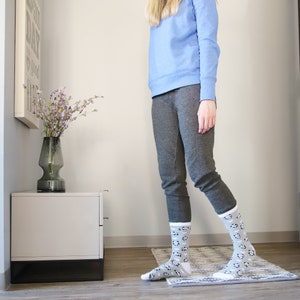 K&ouml;nnte beinhalten: Eine Frau tr&auml;gt ein hellblaues Sweatshirt und graue Jogginghosen mit einem wei&szlig;en und grauen Muster auf den Socken. Sie steht auf einem Teppich mit einer Vase mit lila Blumen im Hintergrund.
