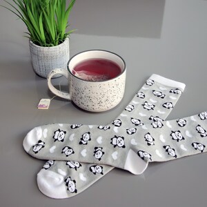 K&ouml;nnte beinhalten: Ein Paar graue Socken mit wei&szlig;em Besatz und einem Pandab&auml;ren-Druck. Im Hintergrund befinden sich eine gesprenkelte Tasse mit einem Teebeutel und eine Topfpflanze.