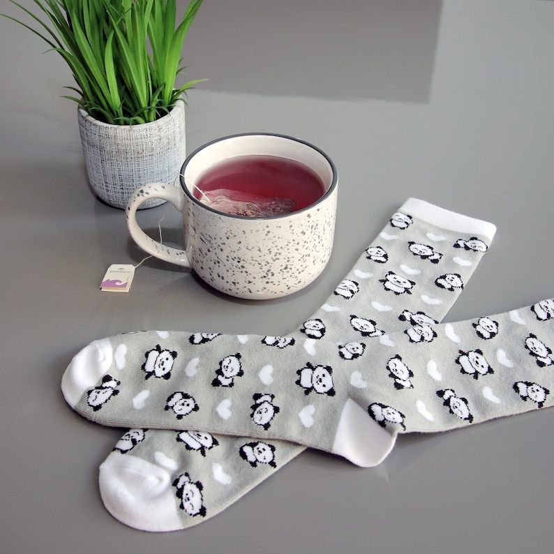 K&ouml;nnte beinhalten: Ein Paar graue Socken mit wei&szlig;em Besatz und einem Pandab&auml;ren-Druck. Im Hintergrund befinden sich eine gesprenkelte Tasse mit einem Teebeutel und eine Topfpflanze.
