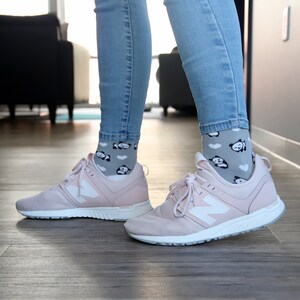 K&ouml;nnte beinhalten: Ein Paar rosa Sneaker mit wei&szlig;en Schn&uuml;rsenkeln und einem wei&szlig;en New Balance Logo. Die Sneaker werden mit grauen Socken mit Panda-Motiv getragen.
