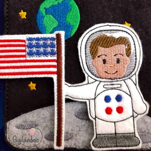 Könnte beinhalten: Ein gestickter Filz-Patch, der einen Astronauten zeigt, der auf dem Mond steht, mit einer amerikanischen Flagge, die in die Mondoberfläche gesteckt ist. Der Astronaut trägt einen weißen Raumanzug mit einem blauen und roten Patch auf der Brust. Der Mond ist grau und der Hintergrund ist dunkelblau mit Sternen.