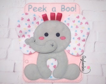 Puede incluir: Un tablero de fieltro rosa con el texto "Peek a Boo!" y un elefante de fieltro gris con patrones de corazones rosas y azules en sus orejas. El elefante tiene un lazo rojo en la cabeza.