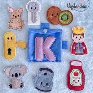 May include: A blue felt pouch with a large purple letter K, surrounded by felt appliques of a kangaroo, key, kiwi, door handle, king, koala, kettle, and a bottle of ketchup with the text Ketchup printed on the front of the bottle.