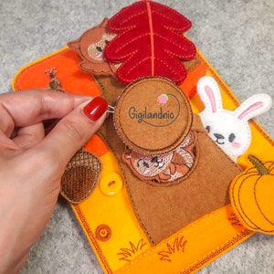 May include: A felt quiet book with a brown tree trunk, a red leaf, a brown squirrel, a white bunny, a brown acorn, a yellow button, and an orange pumpkin. The book has the text "Gigilandnic" on a brown circle.