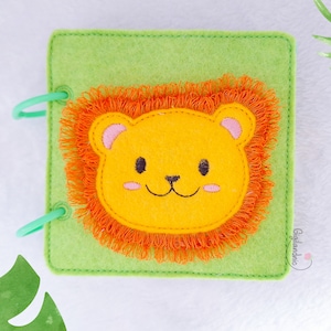 May include: A handmade felt book with a lion design. The cover is light green, with a yellow lion face and orange mane. The lion has pink ears and a smiling face. The book is bound with two green plastic rings.