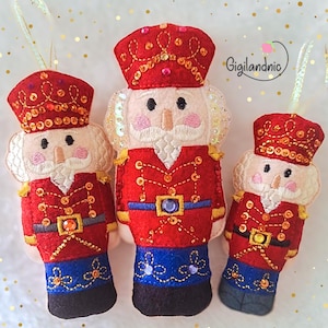 Peut inclure: Trois décorations de casse-noisettes en feutre faits à la main. Chaque décoration présente une veste rouge avec des bordures dorées, un pantalon bleu et un chapeau rouge. Les casse-noisettes ont des cheveux et des barbes blancs, et sont décorés de paillettes et de perles. Ils sont conçus pour être suspendus.