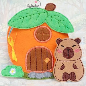 Puede incluir: Una casa de fieltro naranja con hojas verdes y una puerta marrón, junto a un oso de fieltro marrón con mejillas rosas. Un pequeño arbusto de fieltro verde con una flor blanca está a la izquierda. El texto "Gigilandnic" es visible.