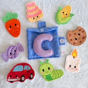 May include: A blue felt pouch with a purple letter C, surrounded by felt shapes including a carrot, a pink cake, a corn cob, a chocolate chip cookie, a purple candy, a red car, a green caterpillar, and a white candle with a yellow flame.