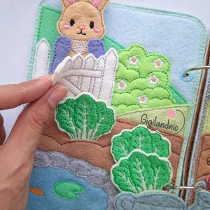 May include: A felt quiet book page featuring a tan rabbit in a blue jacket behind a white picket fence. The scene includes green embroidered leafy vegetables, a blue pond with orange fish, a green lily pad, and a light blue watering can.