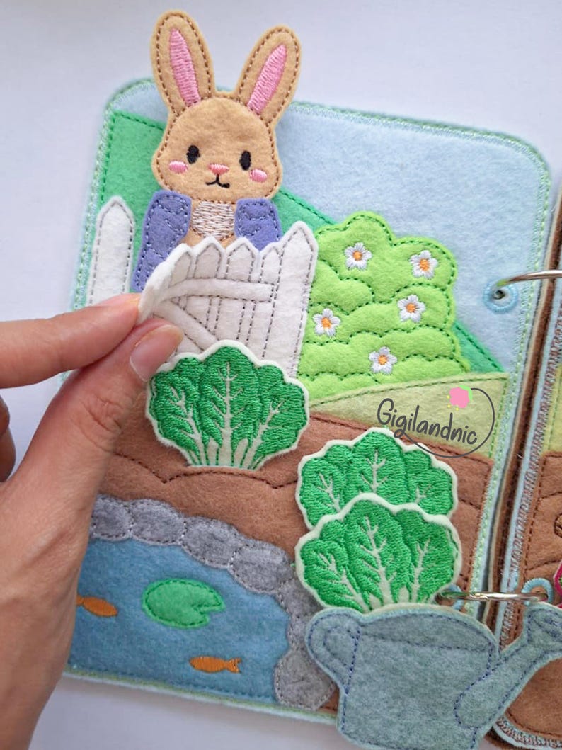 May include: A felt quiet book page featuring a tan rabbit in a blue jacket behind a white picket fence. The scene includes green embroidered leafy vegetables, a blue pond with orange fish, a green lily pad, and a light blue watering can.