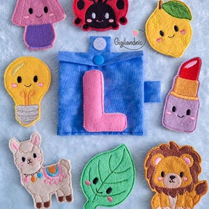 May include: A blue fabric pouch with a pink felt letter L, surrounded by embroidered felt patches including a pink lamp, red ladybug, yellow lemon, yellow lightbulb, red lipstick, beige llama, green leaf, and a brown lion with the text Gigilandnic.
