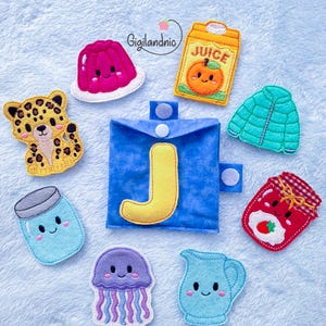 May include: A blue fabric pouch with a yellow letter J on the front, surrounded by felt appliques including a pink gelatin mold, orange juice carton, leopard, teal jacket, blue jar, red strawberry jam jar, purple jellyfish, and a blue pitcher.