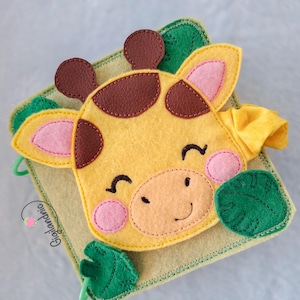 Può includere: Un libro per bambini in feltro fatto a mano con una faccia di giraffa gialla, macchie marroni, guance rosa e un'espressione sorridente. Il libro ha foglie verdi e un elemento in tessuto giallo. Lo sfondo è un quadrato verde chiaro.