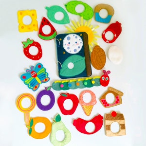 May include: A collection of colorful felt food and nature-themed toys, including a book with a smiling moon, a caterpillar, a butterfly, and various fruits and desserts. The toys are arranged on a white surface.