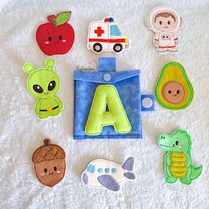May include: A collection of colorful felt appliqués, including an apple, ambulance, astronaut, alien, avocado, acorn, airplane, and a green dinosaur. A blue fabric pouch with a green letter "A" is also present. The appliqués are arranged on a white surface.