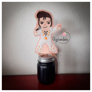 May include: A fabric applique of a cartoon Elvis Presley figure in a white jumpsuit with orange accents, holding a microphone. The applique is attached to a black jar with a silver lid. The background is a neutral wall.