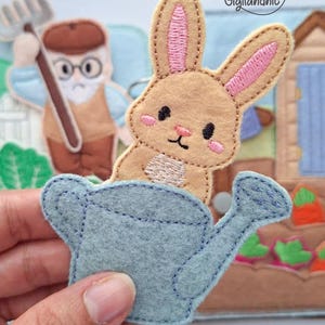 May include: A felt applique featuring a light brown rabbit with pink inner ears and cheeks, peeking out from a light blue watering can. The design includes embroidered details and is part of a larger fabric scene with a gardener figure and text Gigilandnic.
