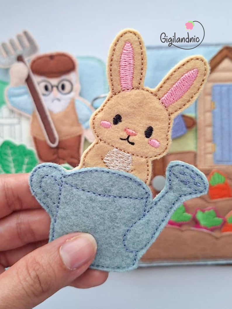 May include: A felt applique featuring a light brown rabbit with pink inner ears and cheeks, peeking out from a light blue watering can. The design includes embroidered details and is part of a larger fabric scene with a gardener figure and text Gigilandnic.